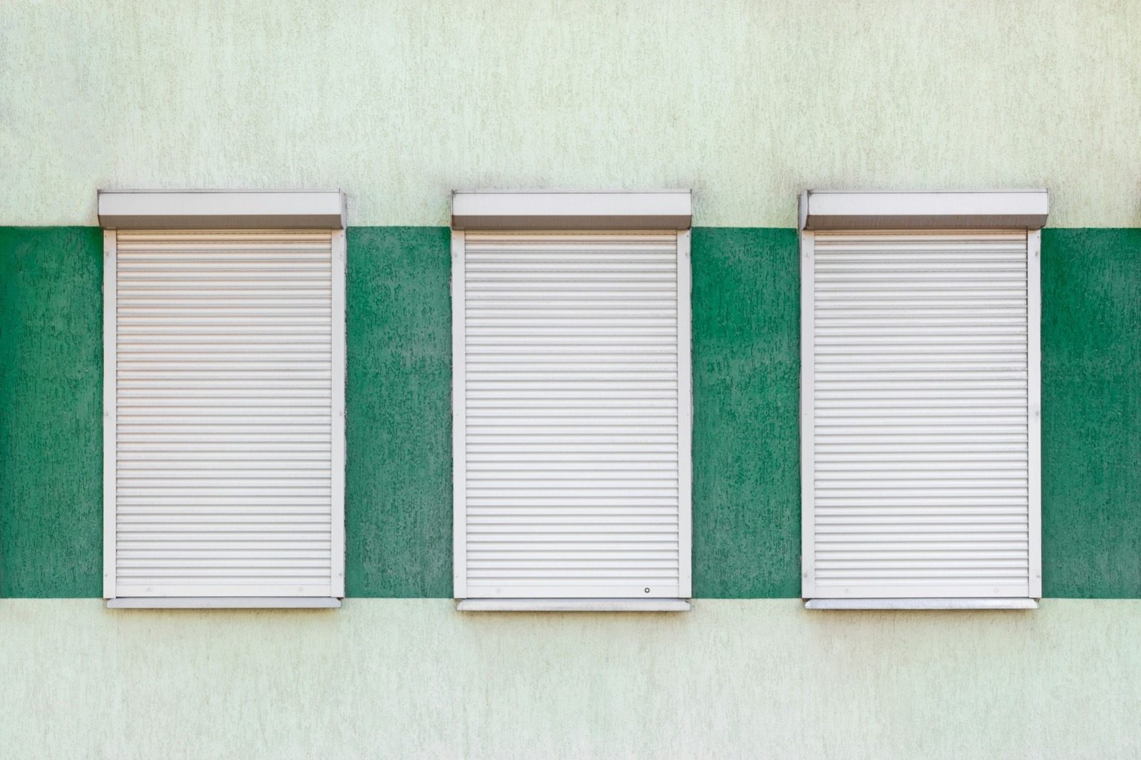 Drei Fenster mit geschlossenen weißen Rollläden an einer grünen Fassade